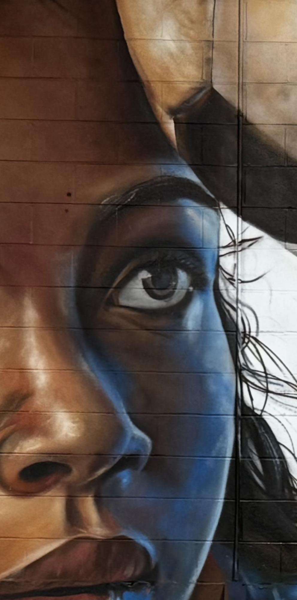 Close-up of a nea.graff mural: a woman's eye and face painted in warm browns and electric blue on a cinder-block wall