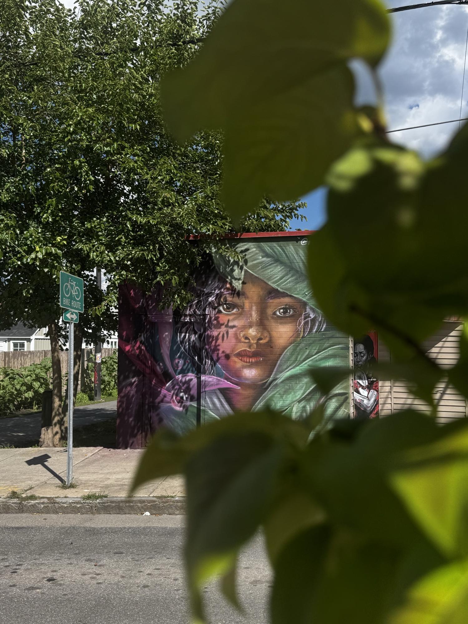 Street mural of a girl's face framed by tropical leaves, seen through real foliage