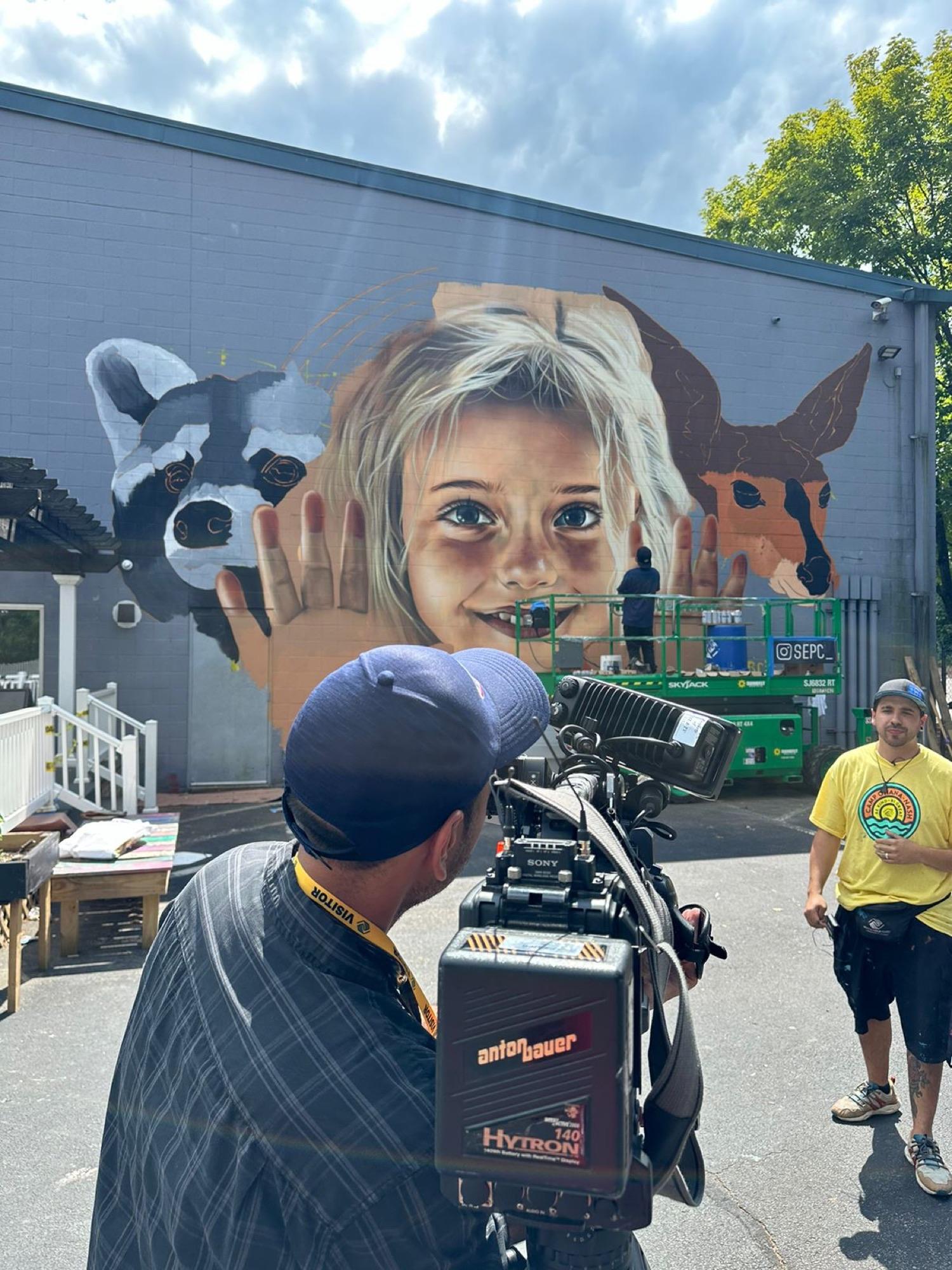 Exterior mural of a child with a raccoon and a fox, being filmed by a camera crew