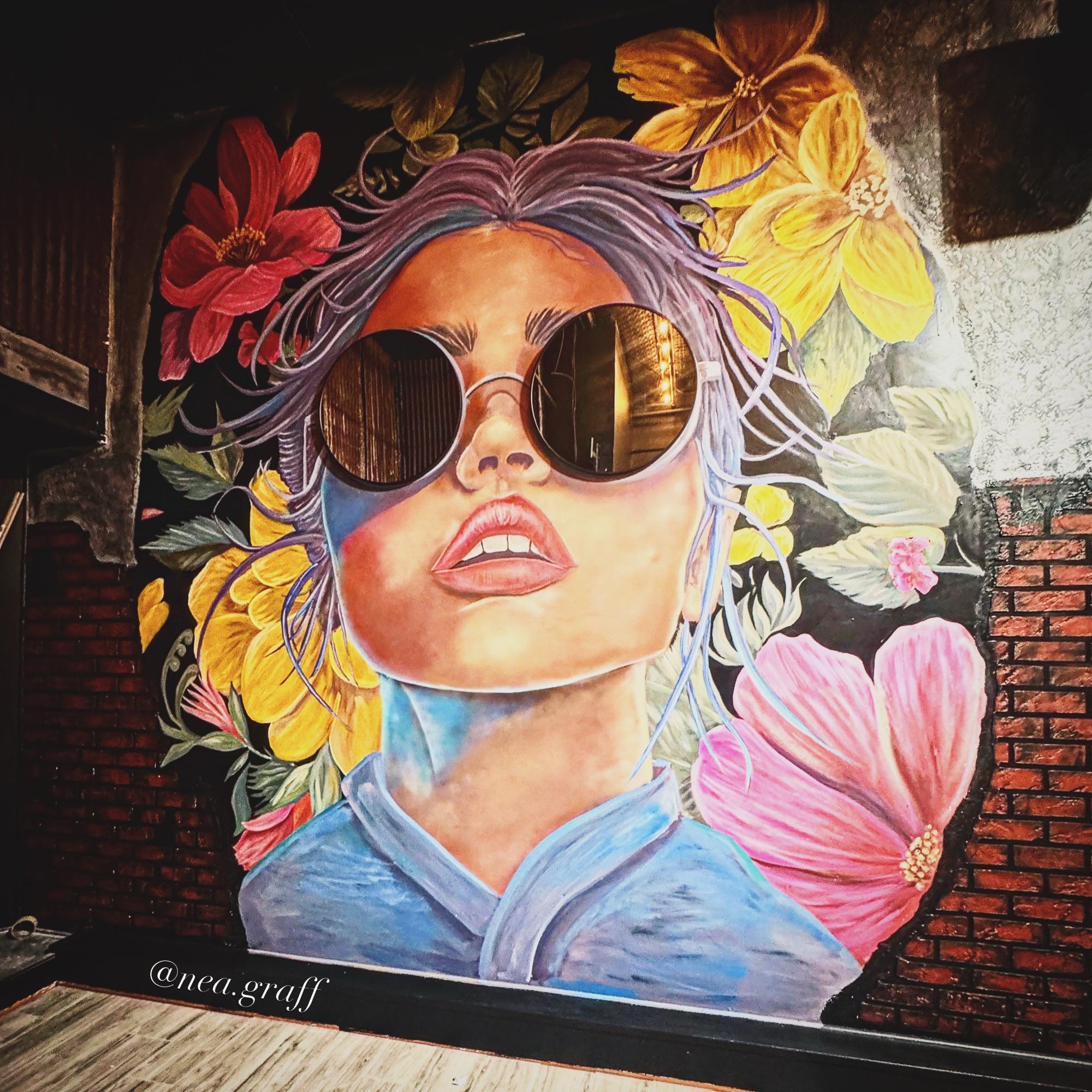 Indoor mural of a woman in round sunglasses framed by giant flowers on a brick wall
